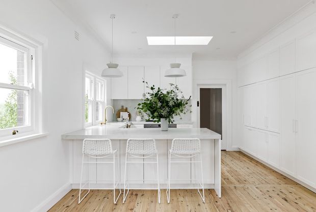 Bright, predominantly white kitchen with a large island and three woven bar stools. The room features white shaker-style cabinetry, white countertops and backsplash, pendant lighting, timber floorboards, a skylight, and abundant natural light from multiple windows. A vase of greenery sits on the island.