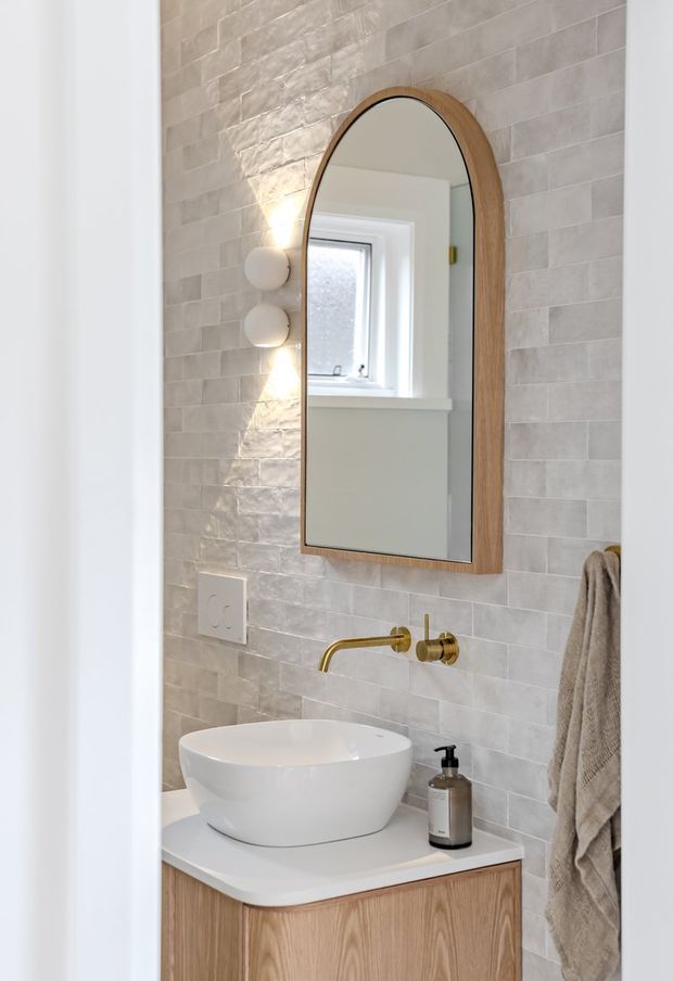 A compact contemporary bathroom vanity viewed through a doorway, featuring a white vessel basin on a light timber vanity, a wall-mounted brushed brass tap, pale textured rectangular wall tiles, and an arched timber-framed mirror with integrated spherical wall lights. A small frosted window is reflected in the mirror.