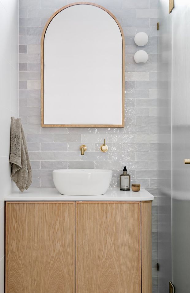 A compact, refined bathroom featuring a pale grey glazed rectangular tile wall, a large arched mirror with a light timber frame, a white vessel basin on a white benchtop, and a light oak vanity with flat-panel cabinetry. Brushed gold tapware and wall lights add subtle warmth, while a textured towel and dark shower glazing are visible at the sides.