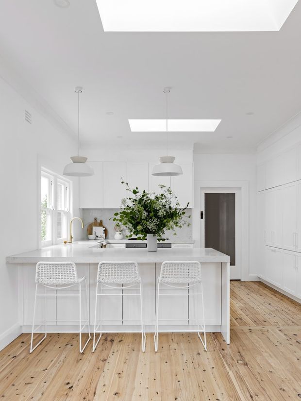 Bright, predominantly white kitchen with a large central island, three woven-back bar stools, white cabinetry, a pale stone benchtop, integrated appliances, pendant lights, skylights, and natural timber floorboards. A floral arrangement sits on the island, with a doorway and full-height cabinetry visible to the right.