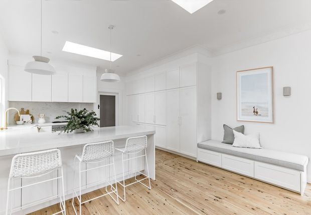 Bright white open-plan kitchen and living area with extensive built-in cabinetry, a large white island with woven bar stools, pale timber flooring, and a built-in window seat beneath framed artwork. Skylights and pendant lights create an airy, refined interior.