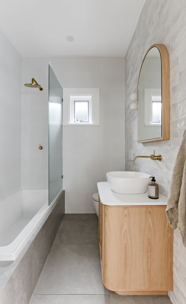 A compact, bright contemporary bathroom with a white bathtub and frameless glass shower screen, grey floor and bath surround tiles, a wall-mounted light timber vanity with white countertop and vessel basin, brushed brass tapware, and an arched timber-framed mirror. White textured wall tiles and a small frosted window complete the space.
