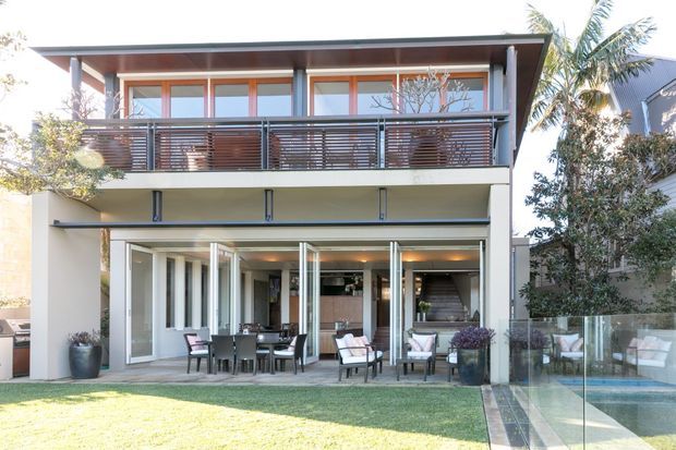 Rear exterior view of a contemporary two-storey house with a broad upper balcony, dark horizontal metal balustrade, timber-framed glazing, and a covered ground-floor outdoor entertaining area with folding glass doors. The property opens onto a lawn and a glass-edged swimming pool, with tropical planting visible around the site.