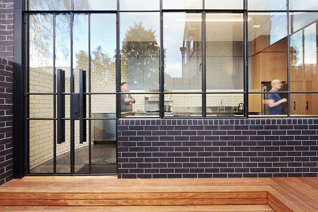Contemporary commercial-style kitchen or workspace viewed from an exterior timber deck through a large black-framed glazed facade. The building features dark brickwork, extensive steel-framed windows and doors, pale brick interior walls, timber cabinetry, and a raised timber deck with integrated stepped seating. People are visible inside, but the architectural fabric and glazing dominate the image.