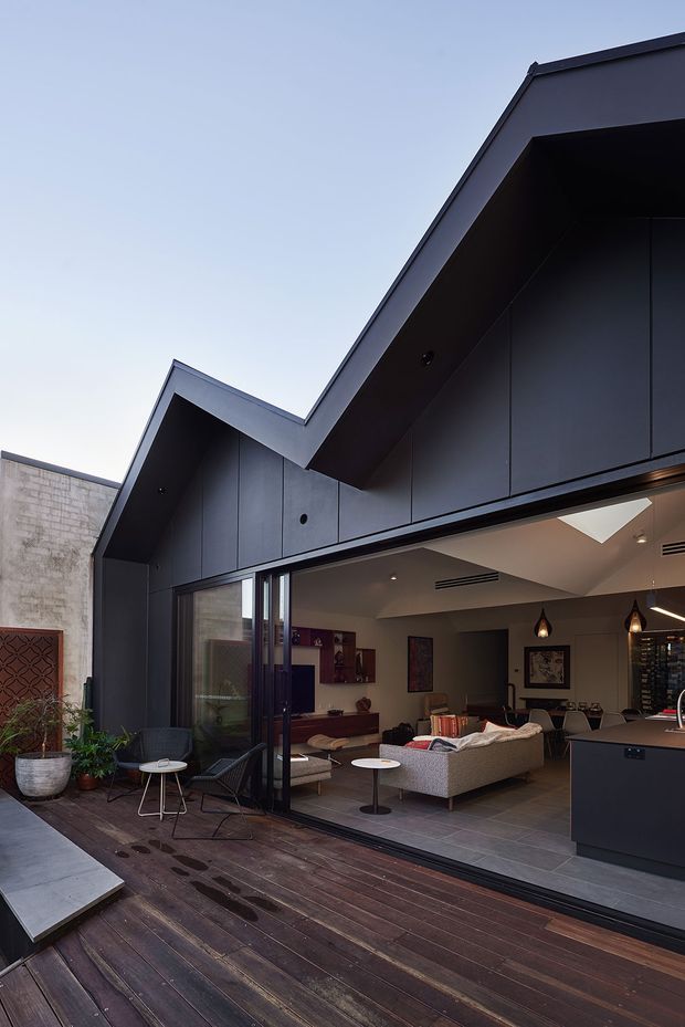 A contemporary dark-clad house photographed from an outdoor timber deck, with a dramatic angular gabled roofline and large open glazing connecting the deck to an open-plan living and dining area. The interior includes a sectional sofa, dining table, pendant lights, artwork and dark kitchen cabinetry. Potted plants and a decorative screen sit along the deck edge.
