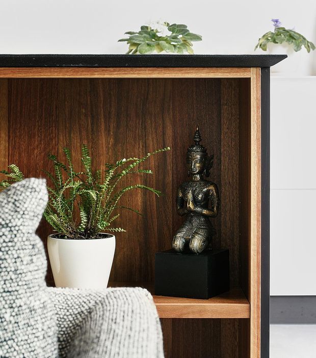 Close-up interior detail of a dark timber built-in shelving unit with open compartments, displaying a potted fern and a dark Buddha sculpture on a black plinth. A textured grey upholstered chair partially obscures the lower left foreground. The joinery has a black top surface, warm timber framing and dark wood backing, with a bright white wall and cabinetry visible to the right.