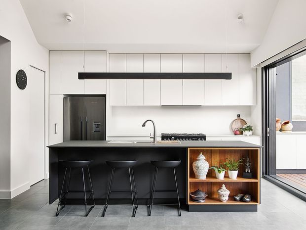 Bright, contemporary kitchen with a long black island and three bar stools, white full-height cabinetry, integrated stainless-steel refrigerator, induction cooktop, and a light benchtop. A timber open display niche with shelving is built into the island end, while large glazed sliding doors open to an exterior balcony or terrace.