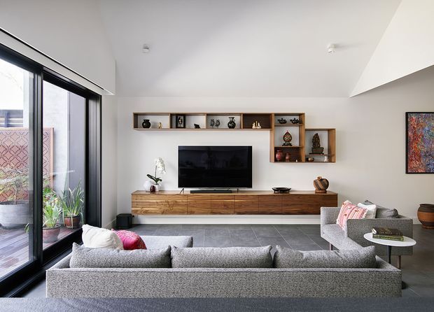 Contemporary living room with a large grey sectional sofa facing a wall-mounted television and long timber media console. Built-in open timber shelving displays decorative objects, while full-height black-framed glazing opens to a planted outdoor area. The room has a white ceiling and walls, dark grey tiled flooring, and a sculptural angled ceiling detail.