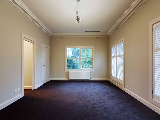 An empty, rectangular formal room with beige painted walls, white decorative crown moulding and trims, dark wall-to-wall carpet, and large white-framed windows fitted with plantation shutters. A central window overlooks greenery, while a radiator sits beneath it. The room has traditional detailing, pendant light fixtures, and multiple door openings.
