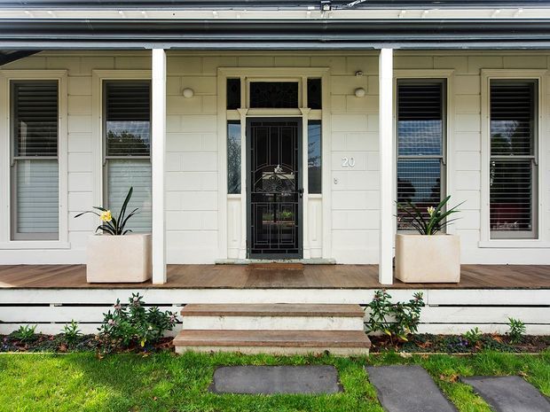 Front elevation of a single-storey traditional residence, centred on a covered entry porch with decorative glazed front door, timber weatherboard cladding, white columns and trim, tall shuttered windows, timber decking, and a short set of steps leading through a landscaped garden.