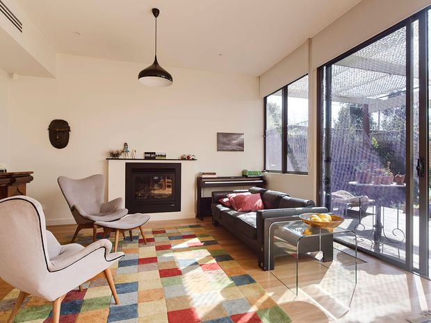 Bright, stylish living room with a fireplace, two sculptural lounge chairs, a dark sofa, colorful patchwork rug, pendant light, and extensive floor-to-ceiling glazing opening to a screened outdoor area. The room has a warm, eclectic mid-century character with abundant natural light.
