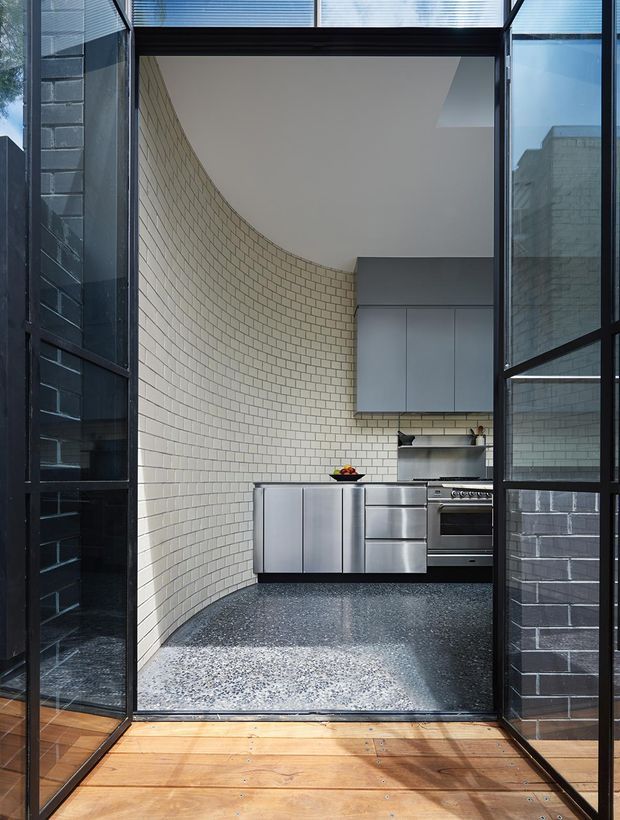 View through a black-framed glazed opening into a compact contemporary kitchen. The kitchen has a curved cream brick-tile wall, stainless-steel cabinetry and appliances, a dark terrazzo-like floor, and a large rooflight or clerestory bringing in natural light. Timber decking or flooring is visible in the foreground, with reflective glazing and dark brickwork framing the opening.