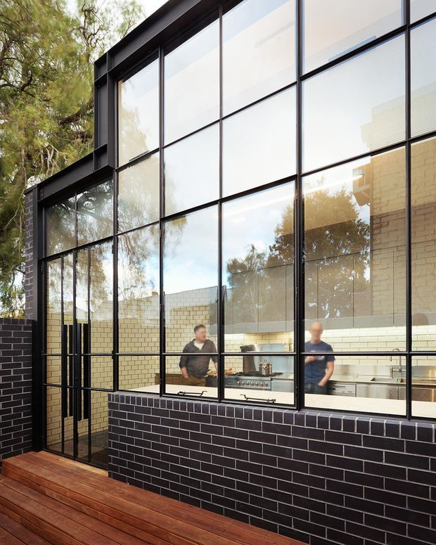 Exterior view of a contemporary building with a dramatic full-height black-framed glazed facade, black brick masonry base, and timber decking. The glazing reveals a bright kitchen interior with pale brick walls, cabinetry, and cooking appliances, while mature trees are reflected in the glass.
