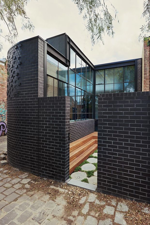 Exterior view of a contemporary urban dwelling or extension, framed by dark charcoal brick boundary walls and a curved brick facade. Large black-framed floor-to-ceiling glazing forms a bright, transparent connection between the interior and a small timber stair and paved entry courtyard. The composition combines strong geometric lines, exposed brickwork, timber decking, and reflective glazing, with neighbouring masonry and mature tree branches visible around the site.
