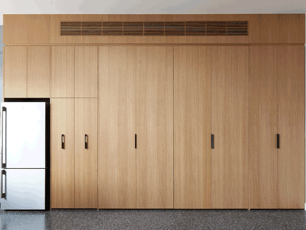 A full-height wall of light timber veneer cabinetry with vertical panelled doors and dark handles. A long linear ventilation grille is integrated near the top, while a white refrigerator is partially visible at the left edge. The cabinetry sits above a dark speckled floor, creating a clean, minimalist built-in storage or kitchen-wall composition.
