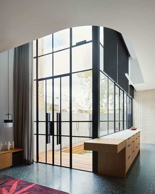 A bright contemporary open-plan interior with a large black-framed steel-look glazed wall and pivot doors opening to a timber deck and courtyard. A long timber kitchen island with integrated drawers occupies the foreground, set against white brick walls and a polished dark terrazzo floor. Floor-to-ceiling curtains, sculptural lighting, and a glimpse of a colourful rug add warmth and contrast. The space has a dramatic curved ceiling opening and abundant natural light.