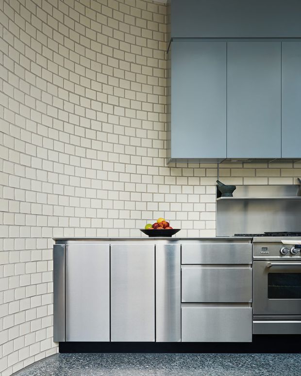Close-up view of a contemporary kitchen with full-height white rectangular tile walls, stainless-steel cabinetry and appliances, a dark terrazzo-like floor, and pale blue-grey overhead cabinets. The compact composition focuses on the cabinetry, benchtop, rangehood area, and freestanding range.