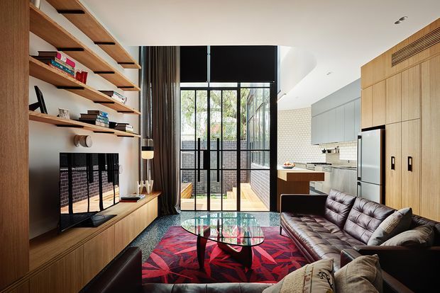 A compact open-plan living room and kitchen with a warm contemporary fit-out. The room features extensive built-in timber shelving and cabinetry, a wall-mounted television, a dark brown leather sofa, a glass coffee table, and a bold red patterned rug. Full-height glazed doors open to a timber deck or courtyard, while the kitchen includes light timber cabinetry, a stainless-steel refrigerator, and a white tiled splashback. High glazing and a bright neutral ceiling create a light-filled yet richly layered interior.