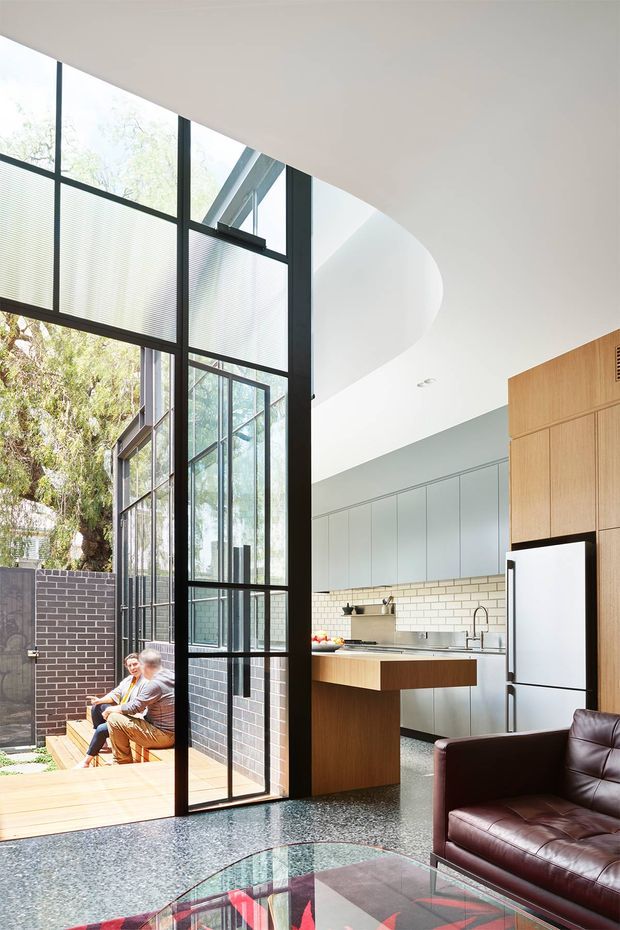 Bright contemporary open-plan interior with a kitchen and living area opening onto a timber deck through dramatic floor-to-ceiling black-framed glazing. The space features a curved white ceiling, polished dark terrazzo-like flooring, pale grey cabinetry, a white subway-tile splashback, timber kitchen joinery, and a deep brown leather sofa. A brick courtyard and mature tree are visible outside.