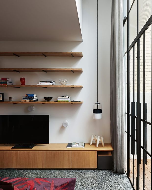 A refined contemporary living room with a warm timber media console, open timber display shelves, and a wall-mounted television. The room has a tall black-framed glazed door or window with sheer curtains, a polished terrazzo floor, and a restrained palette of white, timber, black, and grey. Minimal pendant lighting and carefully arranged books and objects create a calm, curated atmosphere.
