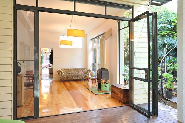 Bright open-plan interior viewed through fully opened black-framed glazed doors, featuring polished timber flooring, cream walls, a patterned sofa, pendant lighting, and a small seating area. The room opens directly onto a timber deck and lush planted outdoor area, with additional glazing and a corridor visible inside.