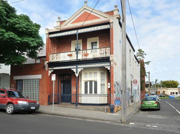 Street-facing view of a narrow two-storey Victorian-era terrace or townhouse with a red brick facade, ornate white decorative trim, lacework verandahs and balustrades, timber-framed windows with shutters, and a steeply pitched gable parapet. The building sits on a dense urban street corner, with adjoining buildings, overhead utility wires, sidewalks, and parked cars visible.
