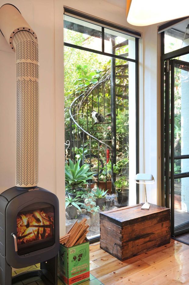 Bright, characterful interior corner with a freestanding black wood-burning stove and tall patterned flue, set beside large black-framed windows overlooking a leafy garden. A rustic wooden chest serves as a side table beneath a slim modern lamp, while timber flooring, potted plants, and a vintage green kindling box add warmth and eclectic detail.