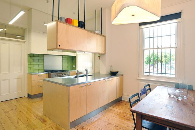Bright open-plan kitchen and dining area with a central timber island, stainless-steel benchtop, pale timber cabinetry, green subway-tile splashback, timber floorboards, and a freestanding dining table beside a large barred window. The kitchen features suspended timber overhead storage and distinctive modern pendant lighting.