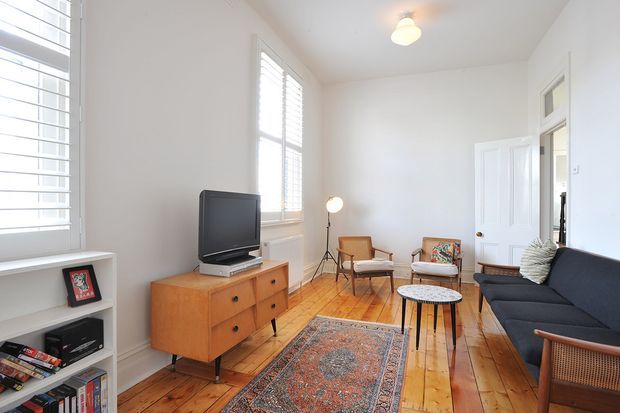 Bright, compact living room with white walls, polished timber floorboards, plantation shutters, a mid-century timber TV console, black sofa, lounge chairs, small coffee tables, and a patterned rug. A doorway opens to an adjacent space, while built-in shelving and a television occupy the left side. The room has a clean, warm, casually styled character with abundant natural light.