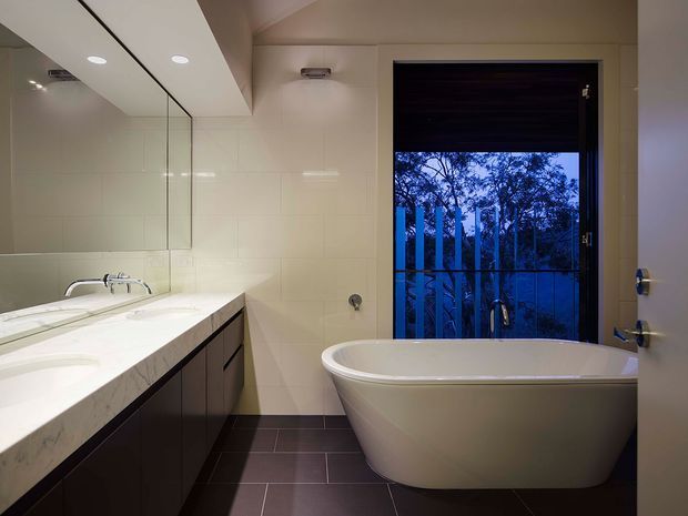 Contemporary bathroom with a freestanding white bathtub positioned beside a full-height window overlooking trees and a dark exterior setting. The room features a long white vanity benchtop with integrated basins, dark fixed cabinetry, large pale wall tiles, dark rectangular floor tiles, a frameless mirrored wall, recessed ceiling lights, and brushed metal tapware.