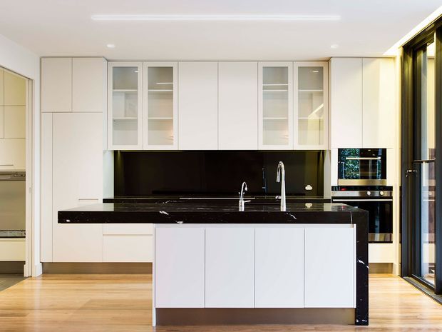 Bright contemporary kitchen with a large white island, integrated cabinetry, glossy black stone benchtops and splashback, glass-front overhead cupboards, and built-in stainless-steel appliances. Warm timber flooring and full-height glazed doors bring natural light into the space.
