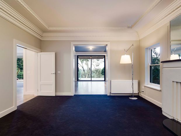 A refined, mostly empty traditional room with pale taupe walls, deep navy carpet, ornate white decorative cornices and ceiling mouldings, tall windows, and a classic white fireplace surround. A doorway opens toward a bright adjoining space, with trees visible outside. A simple floor lamp and radiator are the only furnishings.