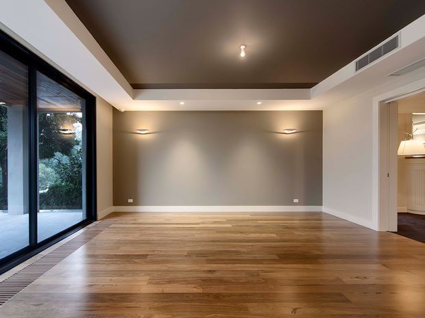 Empty, refined interior room with warm timber flooring, a recessed dark ceiling feature, neutral painted walls, integrated wall lighting, large black-framed glazed doors or windows on the left, and an open doorway leading to another furnished room on the right. The space appears suitable as a living or dining area, with a calm contemporary architectural fit-out.