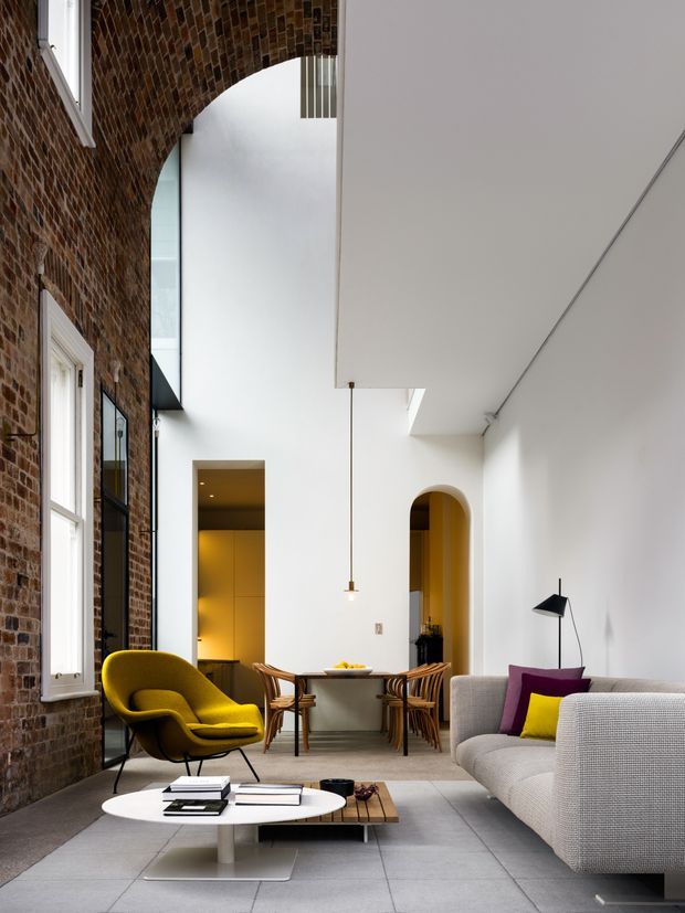 A sophisticated double-height living room combining an exposed red-brick wall with crisp white walls and ceiling. The space features a grey sectional sofa with colourful cushions, a mustard-yellow lounge chair, a low white oval coffee table, timber side table, woven dining chairs, and a sculptural hanging pendant. Arched openings, tall glazing, and a partially visible upper-level void contribute to the dramatic contemporary interior.
