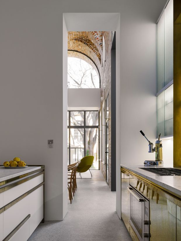 A narrow, refined kitchen viewed toward a tall glazed dining or living area. The kitchen features white and reflective metallic cabinetry, pale stone benchtops, integrated appliances, a dark cooktop, and a polished brass or gold-toned vertical feature at the right. Through the opening, exposed curved brickwork, expansive steel-framed glazing, a sculptural green chair, and dining seating are visible. The space has a high ceiling and strong natural light.