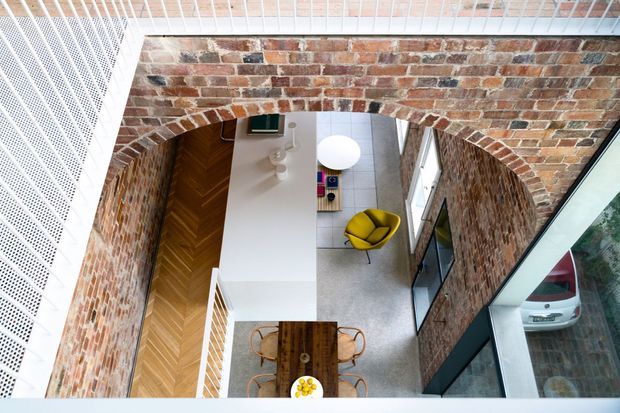 High-angle interior view looking down through a large exposed brick arch into a double-height residential space. The room combines white walls and joinery, warm herringbone timber flooring, a pale hard floor area, a compact dining table, and a bright yellow lounge chair beside extensive glazing. A perforated white metal balustrade or screen is visible at the upper left, and a parked car is seen through the glazing.