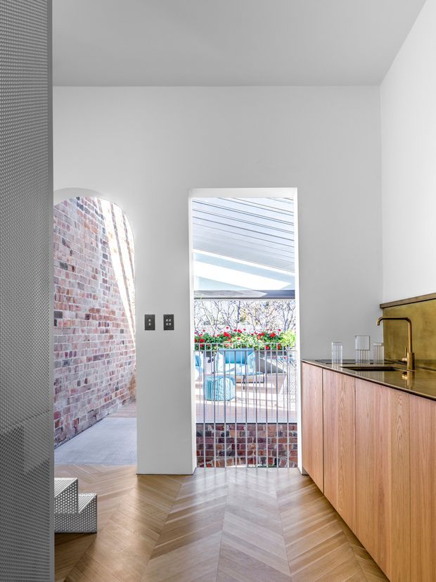 Bright contemporary kitchen interior viewed toward an outdoor courtyard. The space features a warm timber herringbone floor, white walls and ceiling, flat-panel timber cabinetry, a dark benchtop, and a reflective brass-toned splashback. A narrow opening leads to a brick-lined exterior passage, while a glazed doorway opens to a small pool or courtyard area. A perforated metal screen and sculptural stair edge are visible at left.