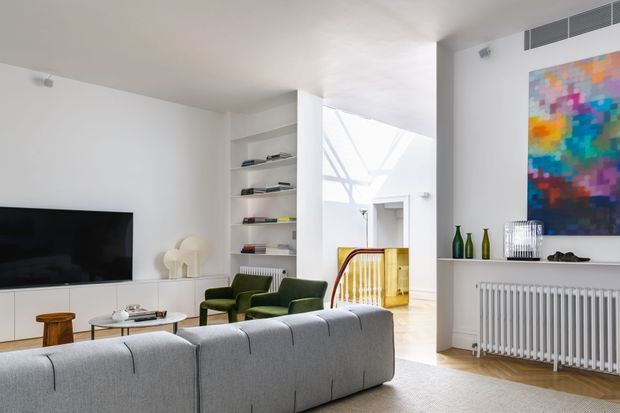 Bright contemporary living room with a large grey sectional sofa in the foreground, green lounge chairs, a low coffee table, wall-mounted television, open shelving, and a white radiator beneath a colourful abstract artwork. A glazed or open stair landing and skylit circulation area are visible beyond the room, with light timber flooring and predominantly white walls and ceiling.