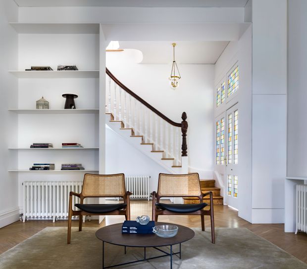 Bright, refined sitting area in a traditional home, centred on two mid-century style cane lounge chairs and a round coffee table. White built-in shelving, ornate stained-glass windows, a timber staircase with turned newel post and dark handrail, vintage radiator, and pendant light create a blend of heritage architectural details and understated contemporary furnishings.