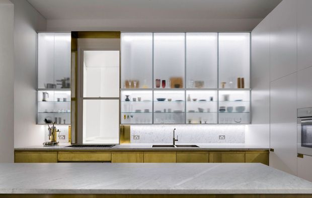 A contemporary galley-style kitchen viewed across a large pale stone island. The space features extensive white handleless cabinetry, illuminated glass-front display cabinets, warm timber-toned lower cabinetry, a white textured backsplash, integrated appliances, and a minimalist sink and tap. The palette is clean and restrained with warm wood accents and subtle brass-toned details.