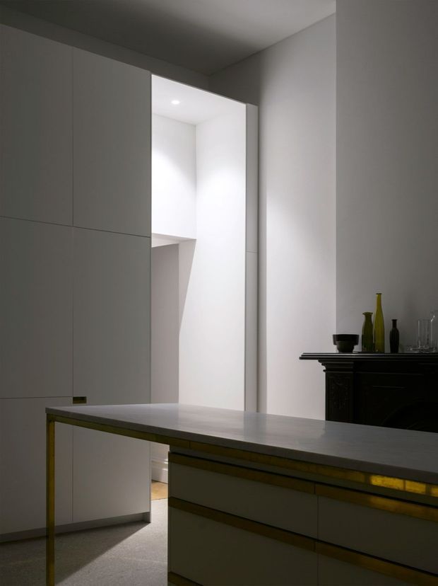 A minimalist, monochromatic interior featuring a large central kitchen island with a pale stone-like benchtop, integrated light-coloured cabinetry, and subtle brass-toned trim. Full-height white built-in joinery lines the left wall. A bright doorway or opening leads to a white-lit space at the rear, while a dark traditional fireplace or mantel with decorative objects contrasts against the otherwise restrained contemporary fit-out. The scene is softly lit with a calm, refined atmosphere.