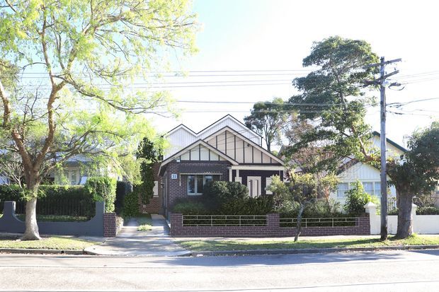 Street-facing view of a compact residential house with a distinctive multi-gabled roofline, dark brick facade, light vertical cladding in the gable ends, white-trimmed windows and entry, and a low brick-and-metal front boundary fence. Mature trees and neighboring homes line the suburban street.