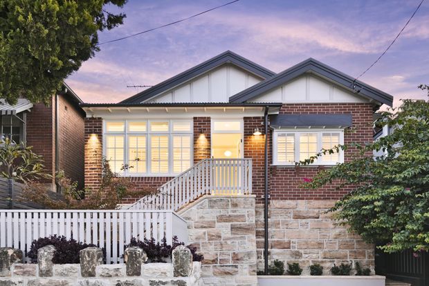 Street-facing view of a single-storey brick home with a layered gabled roof, pale stone retaining wall, white picket fencing and porch stairs. The facade features warm illuminated windows, white trim and a welcoming residential frontage at dusk.