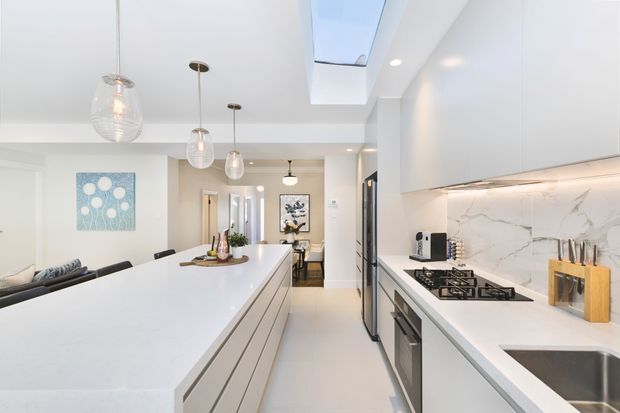 Bright contemporary open-plan kitchen with a large white island, white handleless cabinetry, marble-look splashback, integrated stainless-steel appliances, gas cooktop, pendant lighting and a skylight. The living area is visible beyond the island.