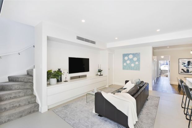 Bright, open-plan contemporary living room with a built-in white media wall and television, grey sectional sofa, patterned area rug, recessed ceiling lights, and a carpeted staircase. The space opens toward a dining or kitchen area in the background and uses a restrained white, grey, and light timber palette.