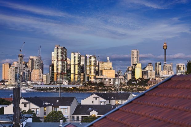 A wide urban skyline view across a waterfront, with dense high-rise development, construction cranes, a prominent communications tower, and low-rise residential houses in the foreground. The image is photographed from an elevated exterior position, with a tiled roof dominating the lower right foreground and a blue evening sky above.