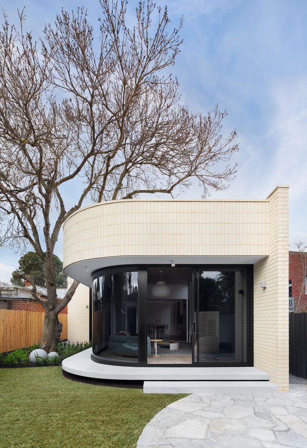 Front exterior view of a refined contemporary brick house extension with a curved cream brick facade, rounded projecting roof canopy, extensive dark-framed glazing, and a small raised entry terrace. A landscaped lawn, mature deciduous tree, timber boundary fence, and irregular pale stone paving are visible. The glazing reveals part of an interior living area.