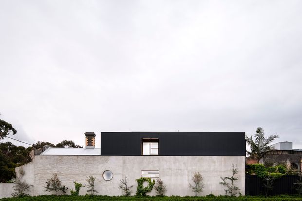Exterior view of a contemporary single-storey home with a long, low rectangular form. The facade combines a pale textured masonry or concrete lower volume with a dark charcoal upper box, a small chimney, simple rectangular glazing, and a circular porthole-style window. Sparse landscaped planting and a palm are visible around the property.