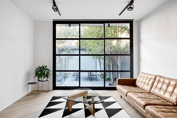 Bright contemporary living room with white walls, pale timber flooring, a large black-framed grid-style glazed door or window opening to a planted courtyard, and a tan tufted leather sofa. A geometric black-and-white rug, sculptural wood-and-glass coffee tables, potted plant, and black track lighting complete the minimalist interior.