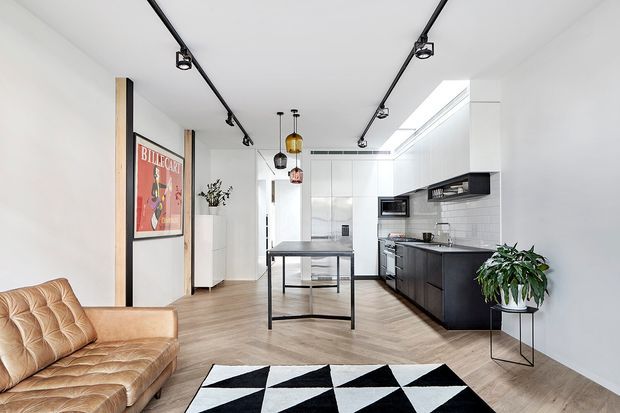 Bright open-plan contemporary interior combining a living area and kitchen. The room has white walls and ceiling, pale timber flooring, a dark fitted kitchen with island, black track lighting, pendant lights, a tan leather sofa, indoor plants, and a bold black-and-white geometric rug. A skylight or roof glazing brightens the kitchen zone.
